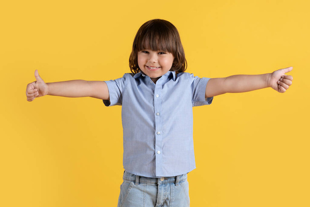 Ειλικρινά παιδιάστικη έγκριση. Studio πορτρέτο του χαριτωμένο χαρούμενο μικρό αγόρι δείχνει αντίχειρες προς τα πάνω χειρονομία με τα δύο απλωμένα χέρια και χαμογελώντας στην κάμερα, πορτοκαλί φόντο - Φωτογραφία, εικόνα