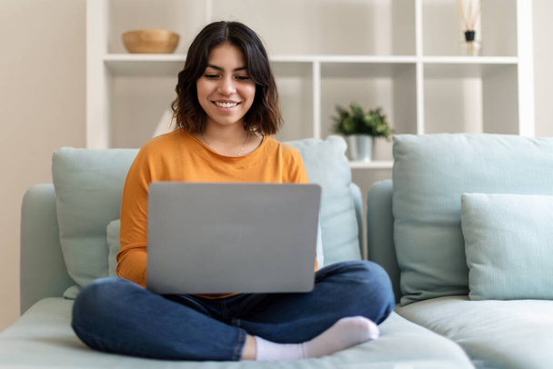 Freelancer árabe sonriente usando el ordenador portátil mientras está sentado en el sofá en casa, feliz joven mujer de Oriente Medio que trabaja en línea en Internet, escribiendo en la computadora, disfrutando de un trabajo remoto, Copiar el espacio - Foto, Imagen