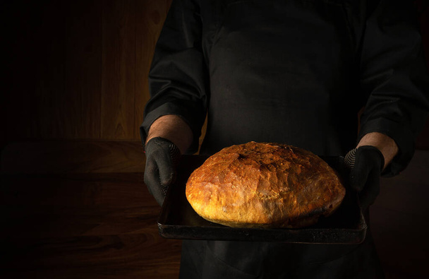 De bakker houdt vers gebakken brood in zijn handen op een pannetje in de bakkerij. Presentatie van bakkerijproducten. Plaats voor menu of recept op donkere achtergrond. - Foto, afbeelding