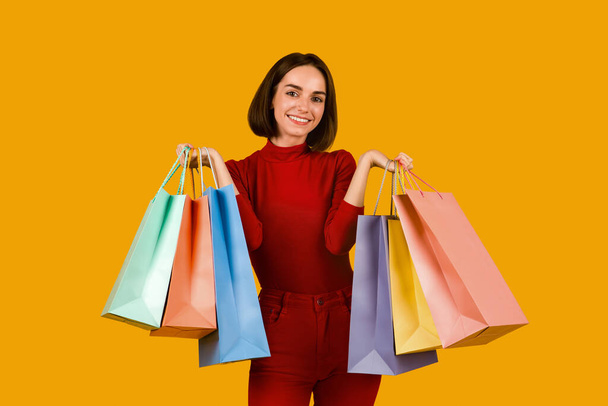 Christamas prodej, nakupování, koncept maloobchodu. Šťastná hezká mladá žena v červené ležérní oblečení s roztomilým účesem drží spoustu barevných nákupních tašek a usmívá se na kameru přes oranžové studio pozadí - Fotografie, Obrázek