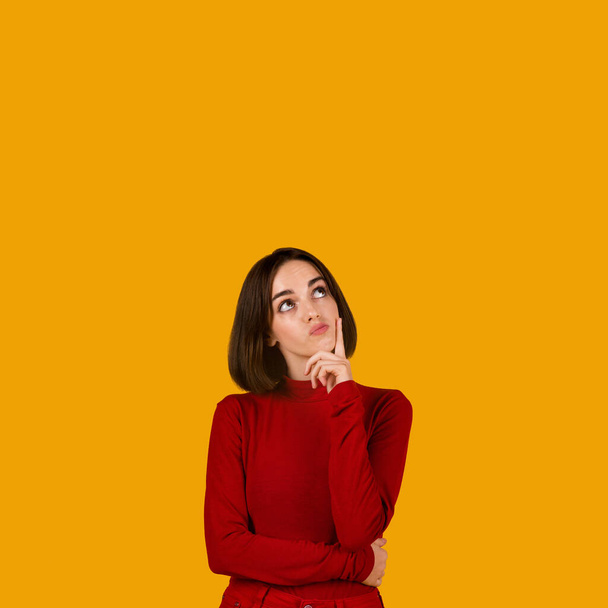 Любопытная задумчивая красивая брюнетка в красном наряде позирует на фоне оранжевой студии, смотрит на пространство для рекламы сверху, трогает подбородок и гримасничает, интересное предложение - Фото, изображение