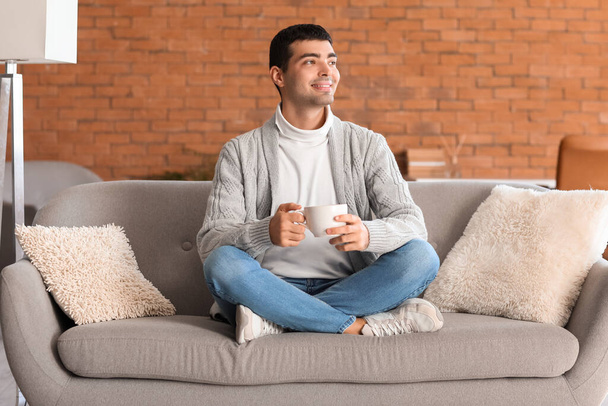 Jeune homme avec une tasse de thé assis sur un canapé gris à la maison - Photo, image