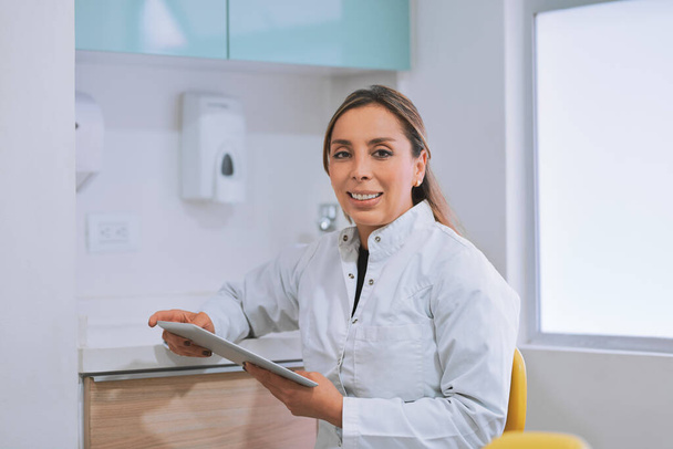 I have been waiting for you. Portrait of a cheerful young female dentist working on her digital tablet while looking at the camera in her office - Foto, Imagen