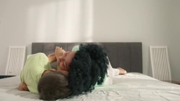 Liefdevolle Afro-Amerikaanse volwassen moeder kietelt kleine schattige kindje zoon. Familie heeft plezier met lachen ontspannen op bed. jong afro amerikaans mam spelen met kleine jongen bonding knuffelen samen - Video