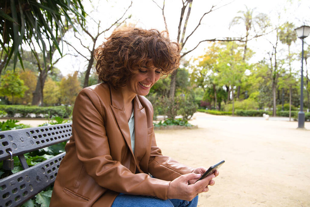 Beautiful woman with curly hair is consulting her mobile phone sitting in the park. The woman is happy while checking her social networks, app, email. Mobile phone concept - Fotografie, Obrázek