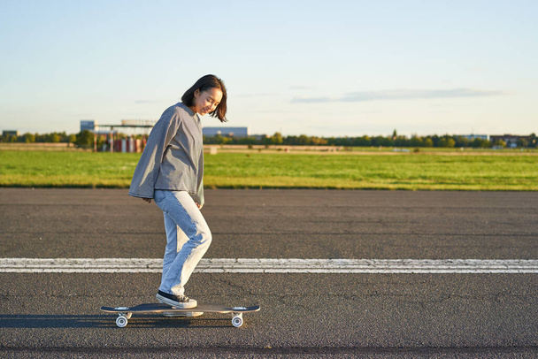 Hermosa chica patinadora asiática montando su longboard en el soleado camino vacío. Joven mujer disfrutando de su paseo en patín sonriendo y riendo. - Foto, Imagen