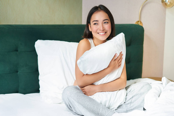 Romantic smiling asian girl lying in her bed, hugging pillow and gazing dreamy, lazy mornings in bedroom. - Photo, Image
