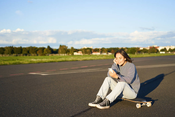 スケートガールは、道路上で彼女のスケートボードに座って、スマートフォンを使用して、モバイルアプリでチャット. - 写真・画像