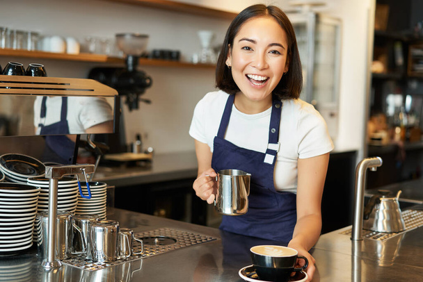 Mooie jonge vrouwelijke barista, het maken van cappuccino, het gieten van gestoomde melk voor latte kunst in beker, geef uw bestelling achter de toonbank. - Foto, afbeelding