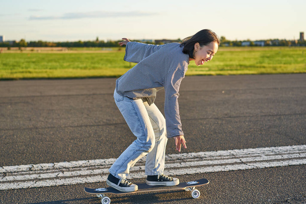 Ευτυχισμένο κορίτσι σκέιτερ ιππασία skateboard της και να διασκεδάσουν σε άδειο δρόμο. Χαμογελαστή γυναίκα απολαμβάνει βόλτα με το καταδρομικό στον ηλιόλουστο δρόμο. - Φωτογραφία, εικόνα