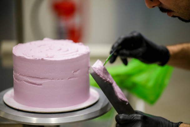 chef pâtissier utilisant spatule et grattoir lissage nivellement couches lilas beurre crème gâteau givré - préparation gâteau de mariage - Photo, image