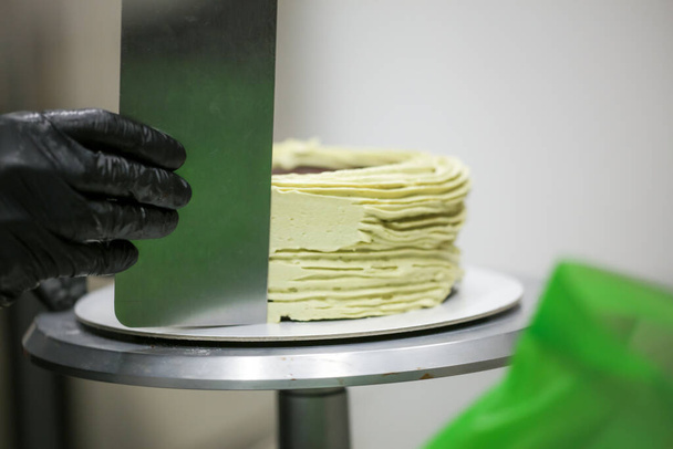 pastelero usando raspador de espátula para suavizar pastel de crema de mantequilla de pistacho verde - preparación de pastel de boda - Foto, Imagen
