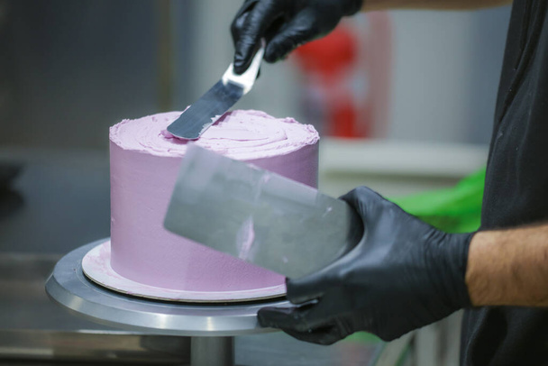 cukiernik stosujący szpachelkę i skrobak wygładzający wygładzający warstwowy maślany krem liliowy ciasto mrożone - przygotowanie tortu weselnego - Zdjęcie, obraz