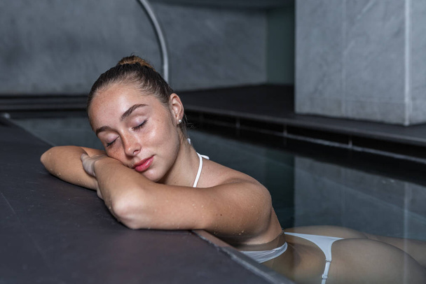 Young female in white bikini closing eyes and holding head on folded arms while chilling in transparent water of clean pool on spa resort - Photo, Image