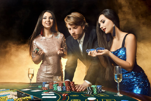 Dvě krásné ženy a mladý muž hrát na pokerovém stole v kasinu, soustředit se na muže a brunetu. Vášeň, karty, žetony, alkohol, kostky, hazard, kasino - to je zábava. Nebezpečná zábava karetní hra pro - Fotografie, Obrázek