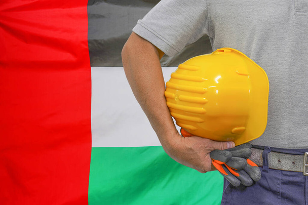Close-up de chapéu duro segurando por trabalhador da construção na bandeira de fundo Emirados Árabes Unidos. Mão de trabalhador com chapéu duro amarelo e luvas. Conceito de trabalhadores industriais em Emirados Árabes Unidos - Foto, Imagem