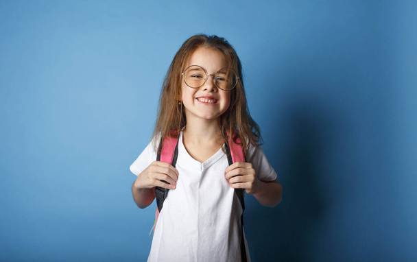 mała uczennica dziewczyna z plecakiem w dużych okularach stoi na niebieskim tle - Zdjęcie, obraz