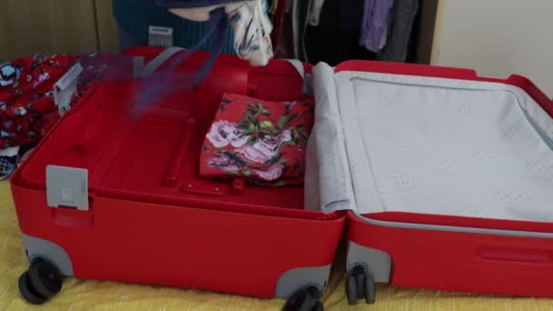 Female hands placing and organizing clothes inside a red suitcase, dresses of different colors and patterns, open on the bed, blue and white colors. Travel concepts - Footage, Video