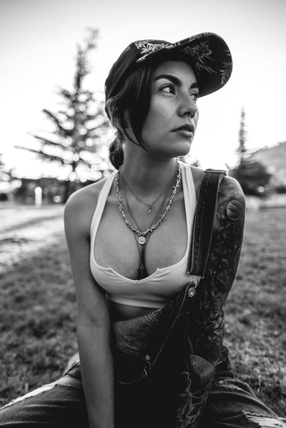 Chica joven grosera, sexy y tatuada con gorra, pantalones vaqueros y top blanco sentado en el parque en un día soleado (en blanco y negro) - Foto, Imagen