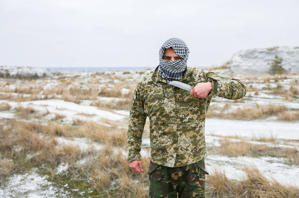 Soldier is standing in the camouflage uniform and checkered keffiyeh shemagh bandana. Man with knife is outdoors in the abandoned deserted place. - Фото, изображение