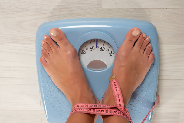 Top view of cropped image of woman feet standing on weigh scales, on the floor. Legs wrapped with a tape measure - Photo, Image