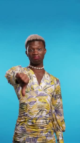 Obrzydliwy Afroamerykanin w stylowej sukience gestykulujący kciuk w dół i patrzący w kamerę na niebieskim tle - Materiał filmowy, wideo