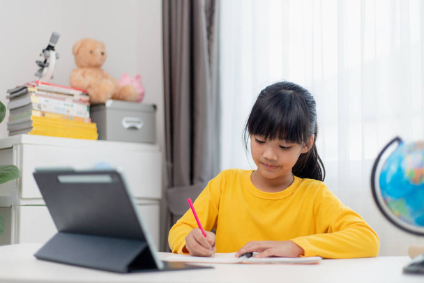 Aasialainen koulutyttö tekee kotiläksynsä digitaalinen tabletti kotona. Lapset käyttävät vempaimia opiskeluun. Koulutus ja etäopetus lapsille. Kotikoulu karanteenin aikana. Pysy kotona. - Valokuva, kuva