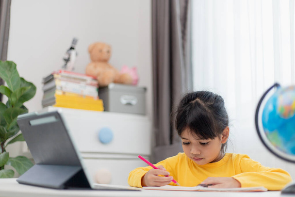 Aasialainen koulutyttö tekee kotiläksynsä digitaalinen tabletti kotona. Lapset käyttävät vempaimia opiskeluun. Koulutus ja etäopetus lapsille. Kotikoulu karanteenin aikana. Pysy kotona. - Valokuva, kuva