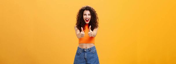 Харизматичные амбициозные и уходящие очаровательные кавказцы. женщина с кудрявой прической и красной помадой, показывая большие пальцы вверх жест в, как или одобрение улыбаясь радостно поддерживая над оранжевой стеной. - Фото, изображение