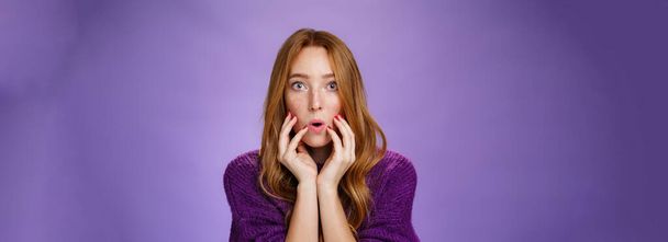 Shocked redhead female friend touching face and open mouth as looking with widen eyes at camera, being overwhelmed with shook and amazement hearing terrible story over purple background. - Photo, Image