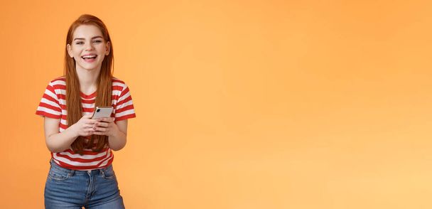 Carefree attractive woman red long hair laughing amused, having fun, chatting friend, hold smartphone, look camera sincere rejoice smile, texting joke, found funny meme internet, orange background. - Photo, Image