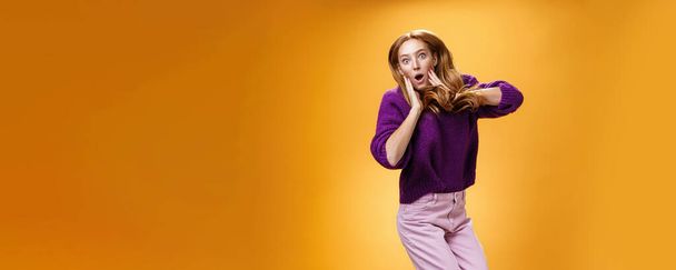Lifestyle. Impressed and surprised amazed good-looking redhead female in purple sweater open mouth astonished and holding hands on cheeks as jumping popping eyes amused over orange background. - Photo, Image