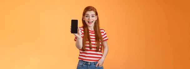 Aufgeregt niedlich ausgehende rothaarige Frau beeindruckt zeigt App, halten Smartphone einführen Gadget-Funktion, Lächeln überrascht, begeistert aussehen wie Anwendung, prahlen Spielstand, orangefarbener Hintergrund. - Foto, Bild