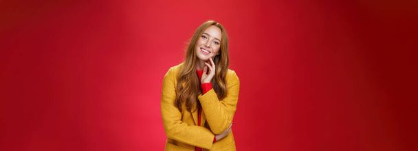 Goed uitziende zorgeloze en gelukkig ontspannen roodharige vriendin in gele stijlvolle jas aanraken gezicht en kantelen hoofd als glimlachen met positieve emoties, poseren vrolijk tegen rode achtergrond. - Foto, afbeelding