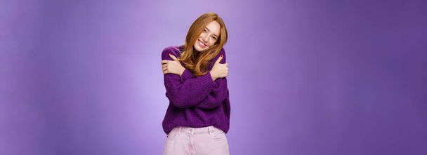 Lifestyle. Silly and cute carefree redhead woman in 20s leaning on shoulder as hugging herself feeling warmth wearing purple sweater smiling broadly in cozy and relaxing atmosphere over violet wall. - Photo, Image