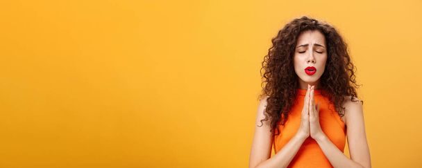 Nervous and concerned woman. with curly hairstyle feeling hopeful praying with closed eyes and frowned eyebrows holding hands in pray near chest hopefully dreaming troubled will solve over orange wall - Photo, Image