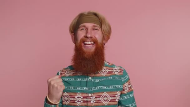 Onnellinen hippi punapää mies kuvio paita huutaa, nostamalla nyrkit ele tein sen, juhlii menestystä, voittaa, syntymäpäivä, arpajaiset tavoite saavutuksia. Hipster kaveri poika sisätiloissa vaaleanpunainen studio tausta - Materiaali, video