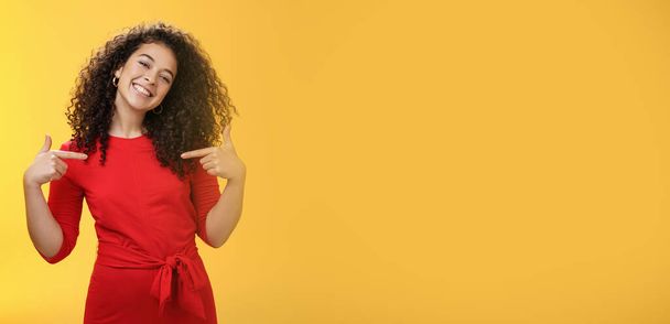 Lebensstil. Stolze und zufriedene ehrgeizige erfolgreiche Studentin im roten Kleid steht zufrieden lächelnd da und zeigt auf sich selbst, als prahle sie glücklich und froh über die gelbe Wand mit ihren eigenen Leistungen. - Foto, Bild
