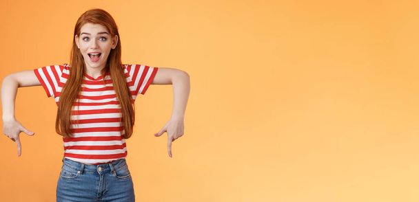 Enthusiastic lively cute redhead woman introduce incredible offer, pointing down, bottom copy space, smiling amazed, look camera, explain you awesome opportunity, feel excitement, orange background. - Photo, Image