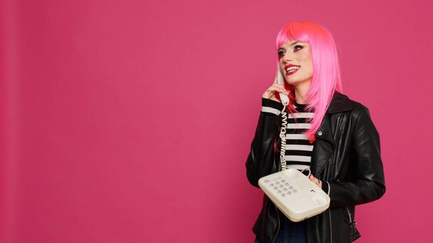 Atrakcyjna kobieta z różową peruką rozmawiająca przez telefon stacjonarny, rozmawiająca na retro linii telefonicznej z kablem w studio. Zabawa przy użyciu telefonu vintage do zdalnej komunikacji, fajna osoba. - Zdjęcie, obraz