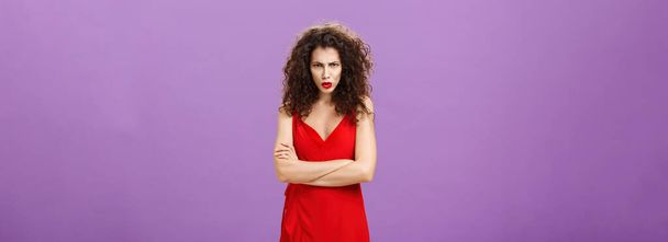 Kırmızılı, zarif kırmızı gece elbisesi giymiş, göğsünü göğsüne dayamış, öfkeyle alnının altından bakan ve eleştirilen kızgın bir kadının stüdyo fotoğrafı. - Fotoğraf, Görsel