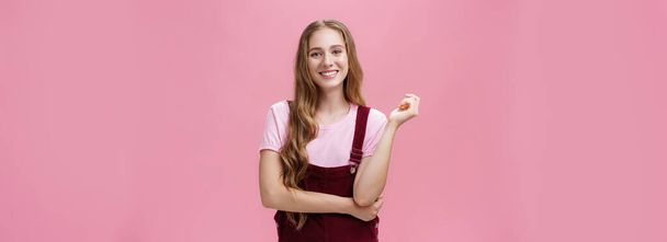 Cute girl in trendy overalls with long wavy hairstyle and light makeup standing in casual pose and smiling cheerfully at camera listening taking part in conversation with pleasant positive attitude - Photo, Image