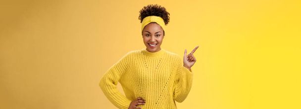 Urocza, szykowna, afrykańsko-amerykańska dziewczyna. w sweter wskazując górny lewy róg stoi pewnie zuchwały energiczny uśmiech żółty tło promujące merch pokazując najlepszy wybór aparat wygląd zabawy. - Zdjęcie, obraz
