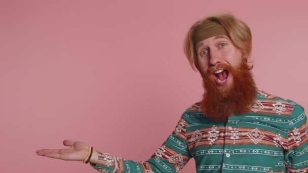 Hippie rudy brodaty mężczyzna we wzorzystej koszuli pokazuje kciuki do góry i wskazując w lewo na pustej przestrzeni. Miejsce na logo reklamowe, baner, tekst. hipster imbir facet chłopiec na różowym tle - Materiał filmowy, wideo