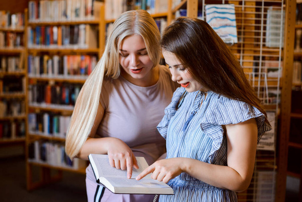 Dos niñas uni estudiantes en la biblioteca discutiendo la trama de una novela, de pie entre estanterías aireando puntos de vista e impresiones sobre el libro - Foto, Imagen