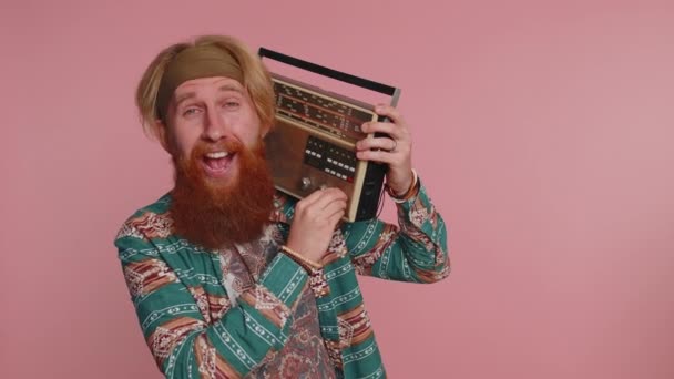 Hippie κοκκινομάλλης άνθρωπος χρησιμοποιώντας ρετρό ταινία πικάπ για να ακούσετε μουσική, ντίσκο χορό του αγαπημένου κομματιού, διασκεδάζοντας, διασκεδάζοντας, οπαδός των vintage τεχνολογιών. Hipster τζίντζερ γενειοφόρος τύπος σε ροζ φόντο - Πλάνα, βίντεο