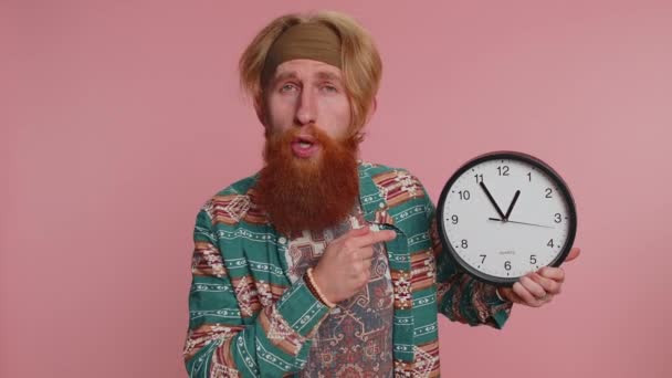 It is your time. Portrait of bearded young hippie man in pattern shirt showing time on clock watch, ok, thumb up, approve, pointing finger at camera. Hipster guy indoors studio shot on pink background - Footage, Video