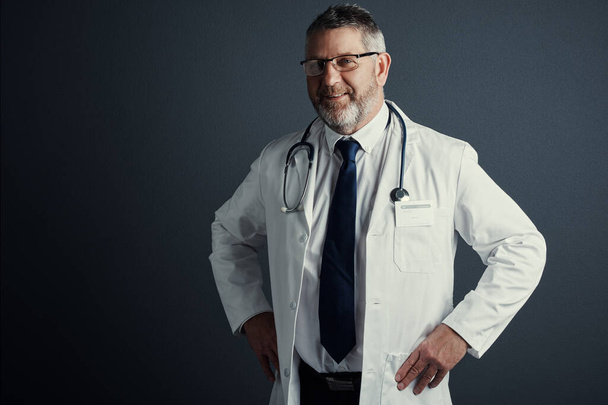 Ik dek je wel. Studio portret van een knappe volwassen mannelijke arts staan met zijn handen op zijn heupen tegen een donkere achtergrond - Foto, afbeelding