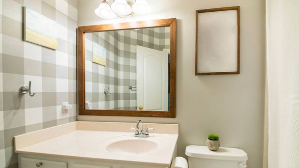 Πανόραμα Μικρό εσωτερικό μπάνιο με καρό ταπετσαρία στα δεξιά. Υπάρχει μια τουαλέτα με γλάστρα και πλαίσιο εικόνας κοντά στον πλαισιωμένο καθρέφτη στα δεξιά πάνω από το νεροχύτη ματαιοδοξίας με λευκά συρτάρια. - Φωτογραφία, εικόνα