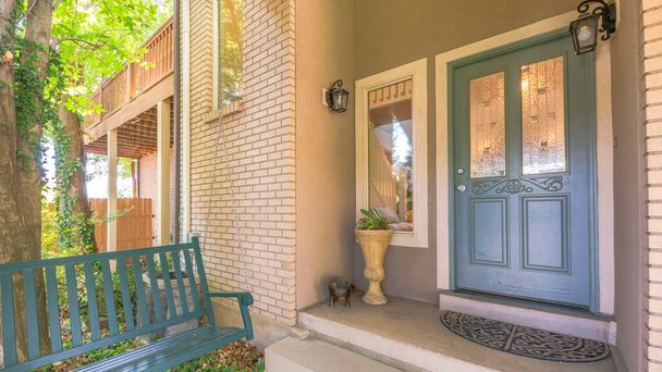 Panorama Gray front door with ornate glass panel near the picture window and wall lamps. Entrance of a house with bricks and hanging bench on the right against the tree, fence and railings of the terrace. - Photo, Image